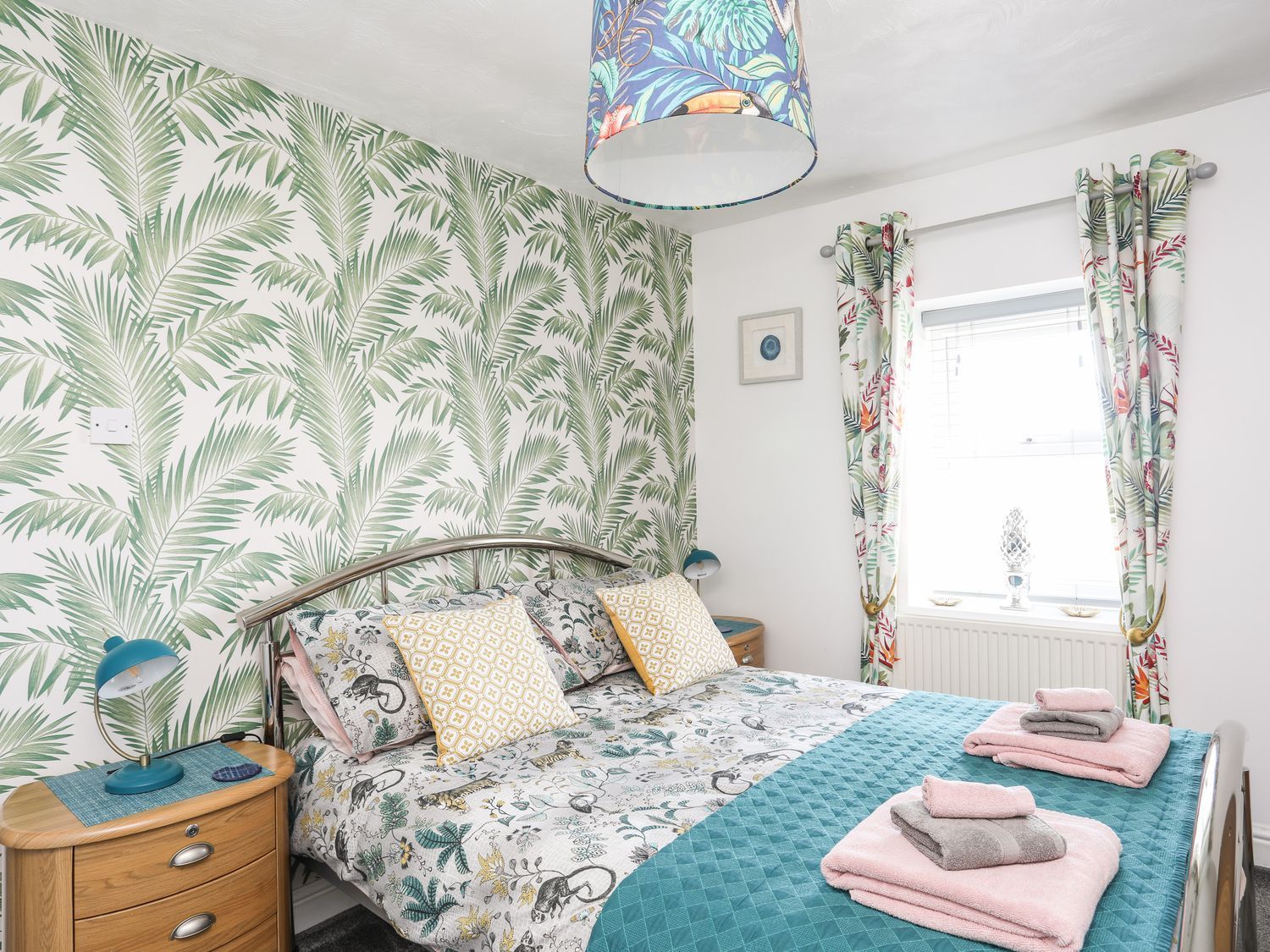 Bright bedroom with tropical wallpaper, floral bedding, teal throw, and a window with patterned curtains