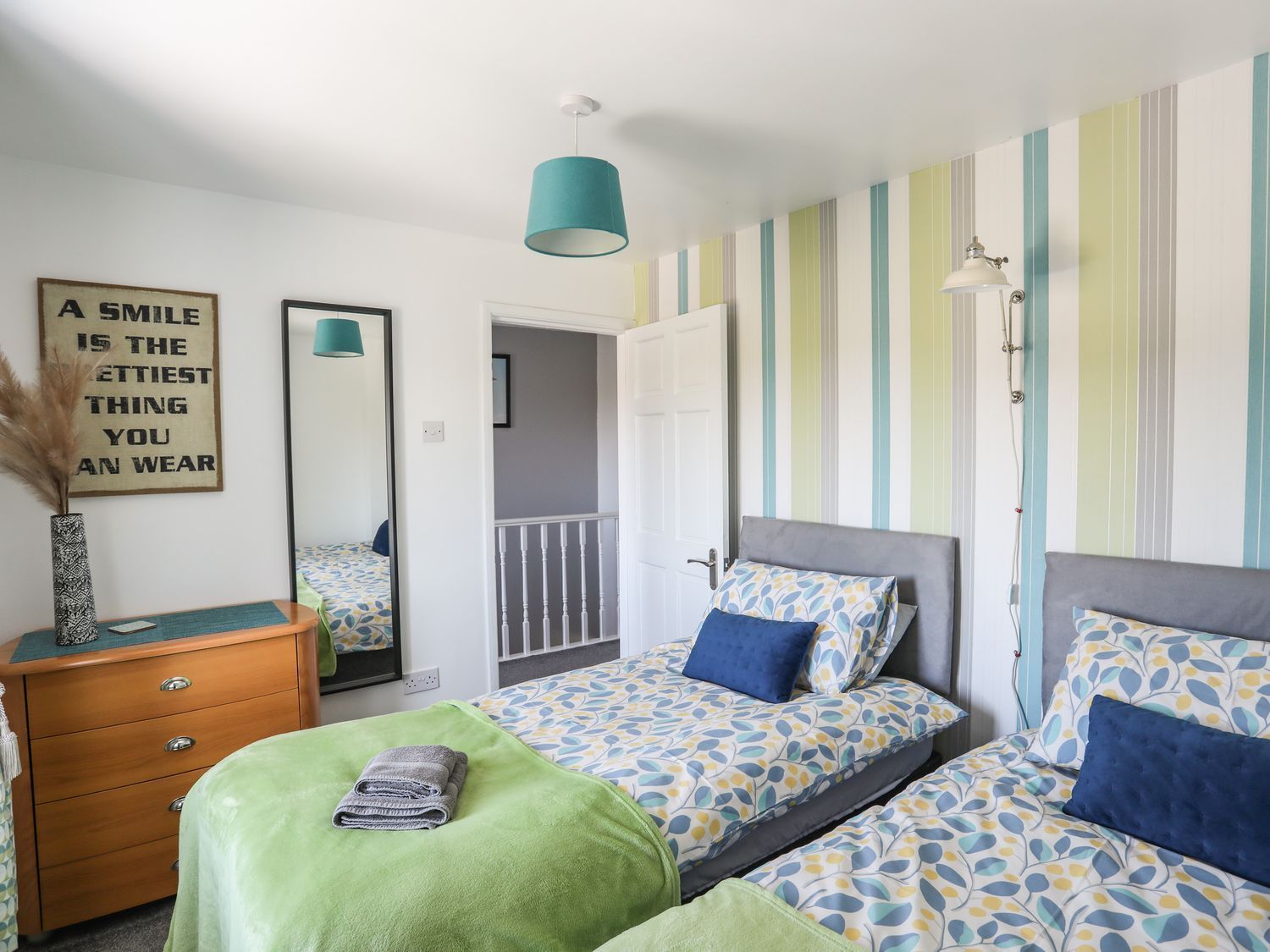 Bright twin bedroom with patterned blue bedding, striped wallpaper, dresser, mirror, and turquoise lamp.