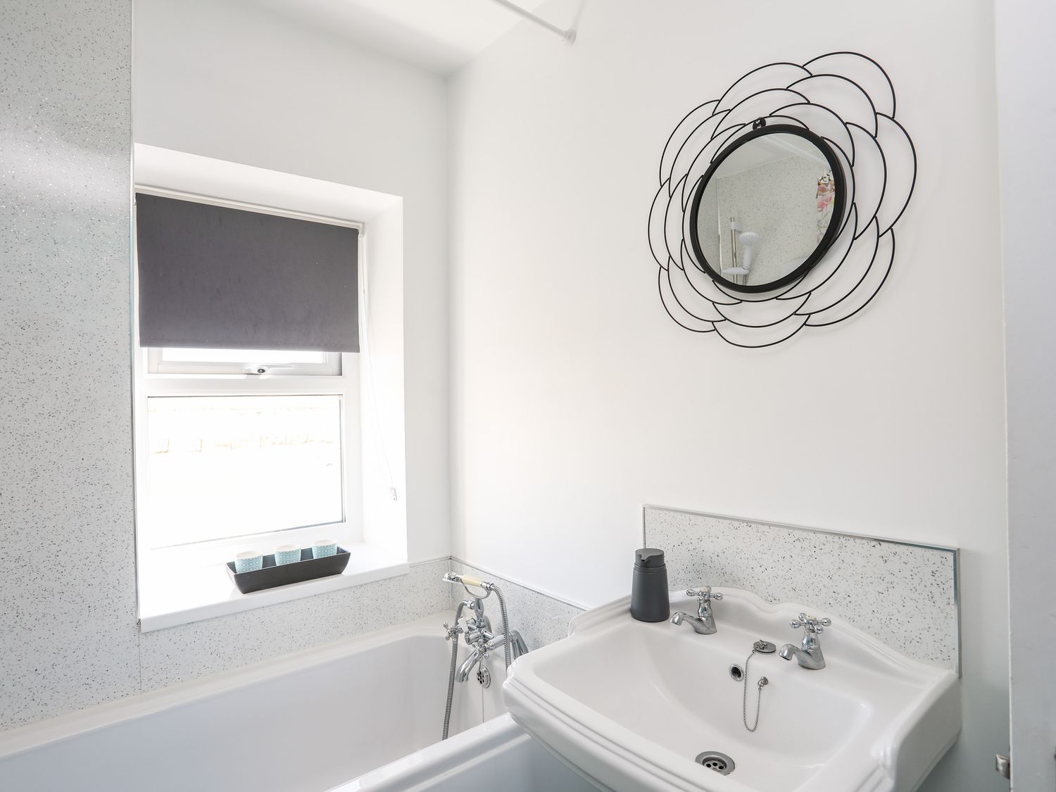 White bathroom with sink, bathtub, window blind, and decorative wall mirror.
