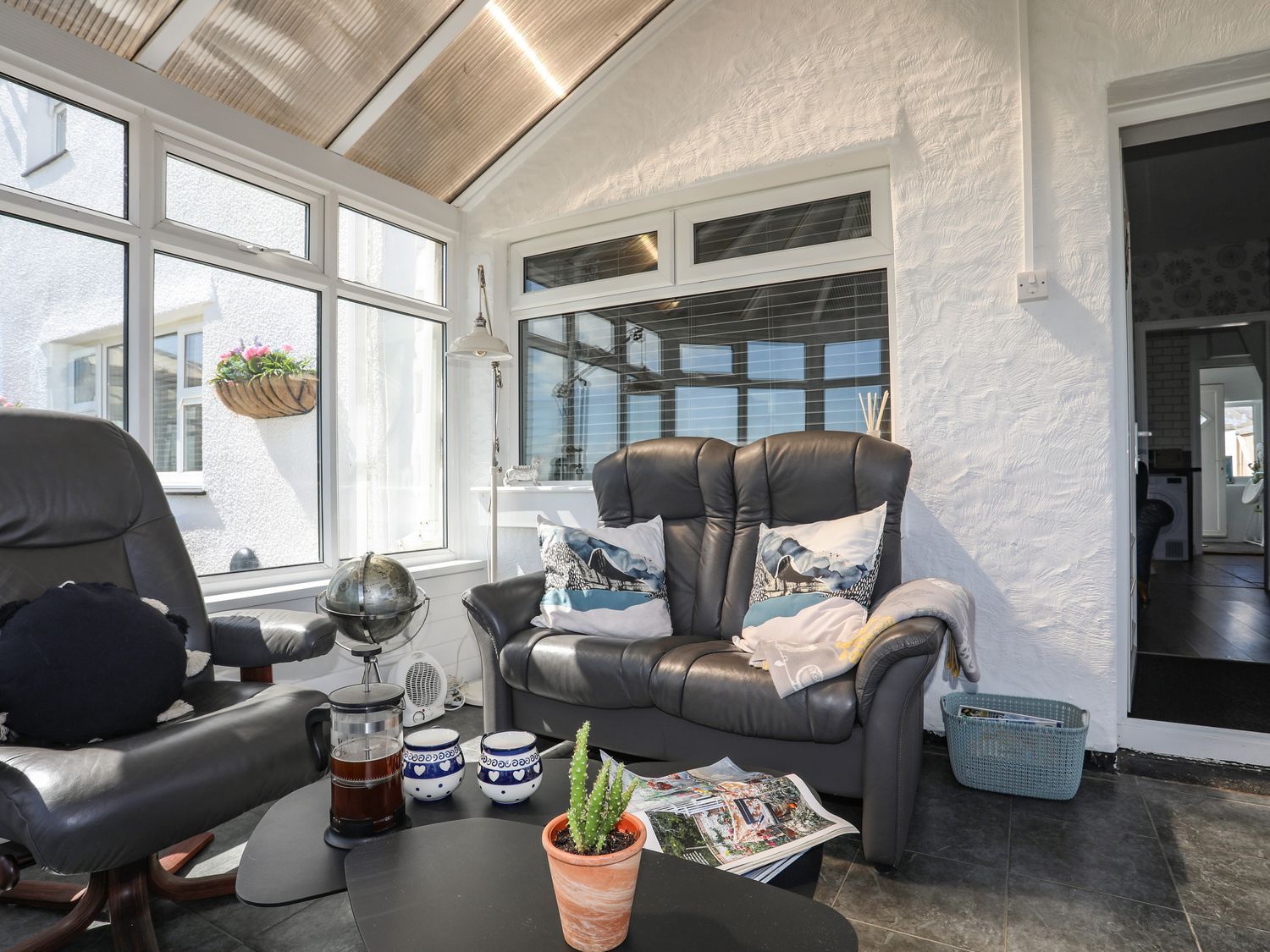 Sunlit enclosed porch with gray recliners, coffee table, hanging plant, and large windows