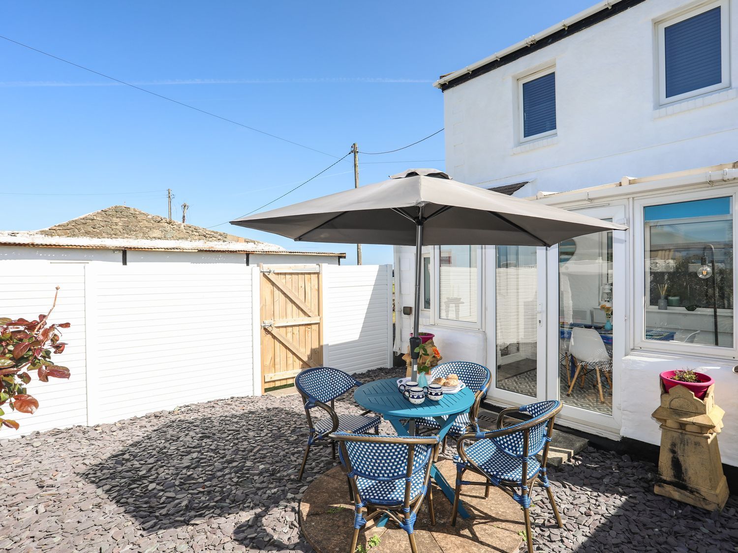 Patio with blue table, chairs, umbrella, potted flowers, and white fence beside a house