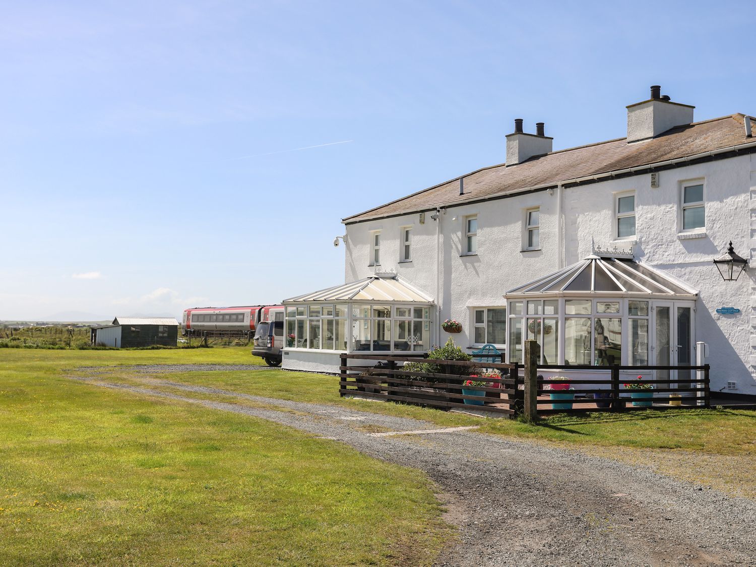 White seaside cottage with glass porch, gravel path, and open grassy field under a clear blue sky