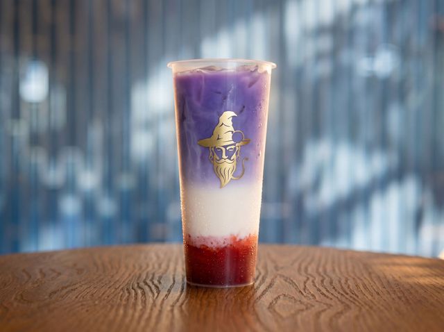 Tall layered purple-to-white-to-red drink with a gold emblem on a wooden table, blurred blue background