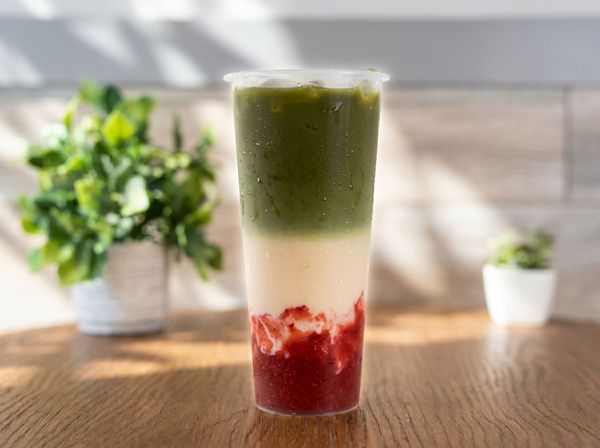 Layered green, cream, and red smoothie in a clear glass on a sunlit wooden table.