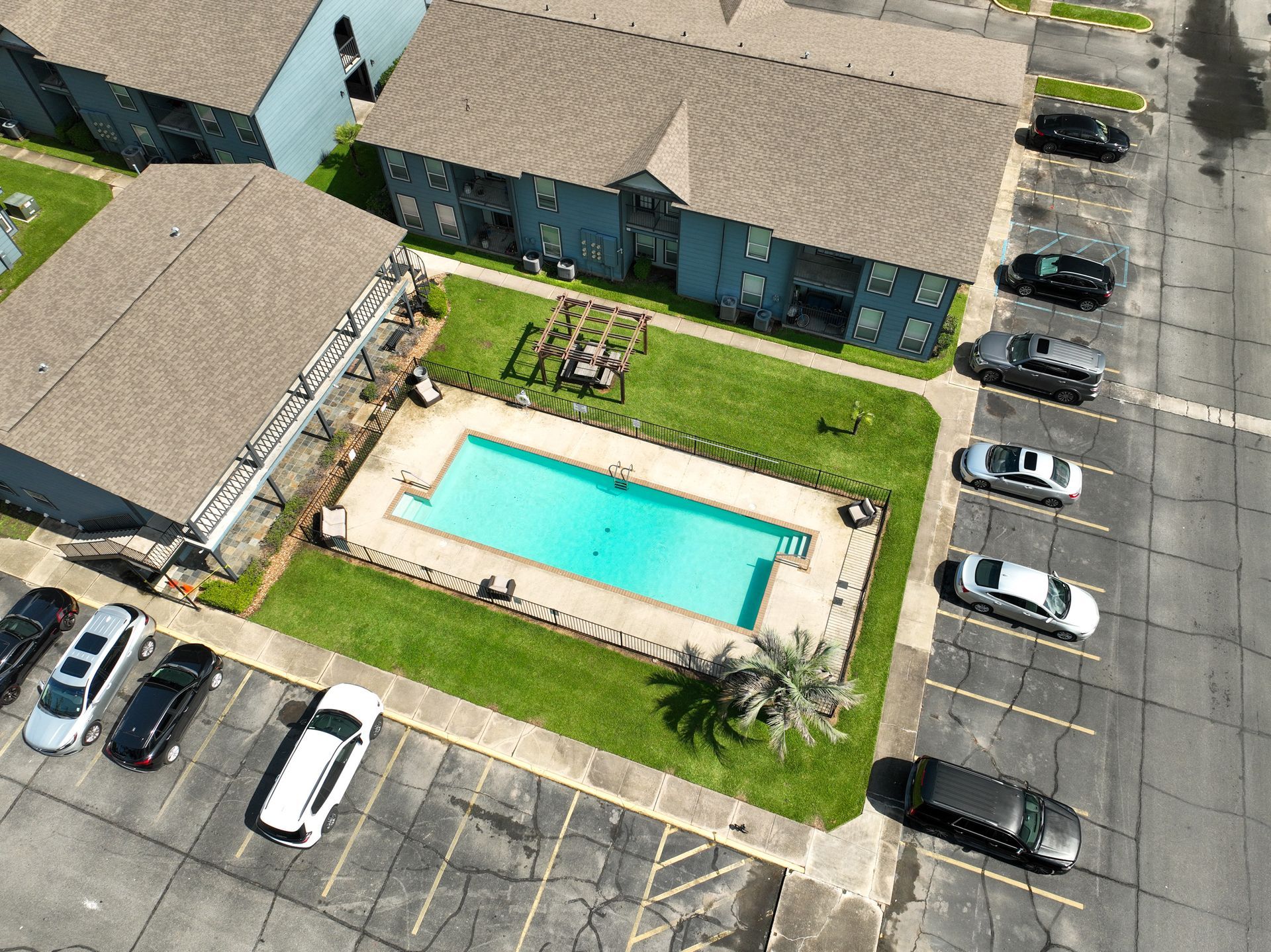 Aerial view of an apartment complex courtyard with a swimming pool, lawn, palm trees, and parked cars