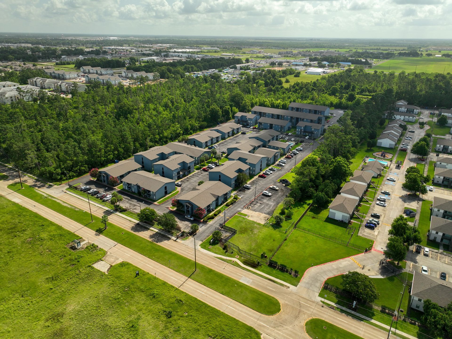 Aerial view of a suburban neighborhood with rows of houses, trees, roads, and green fields beyond.