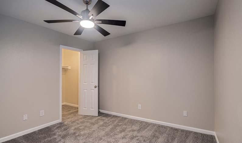 Empty gray bedroom with carpet, ceiling fan, and open door to a lit hallway