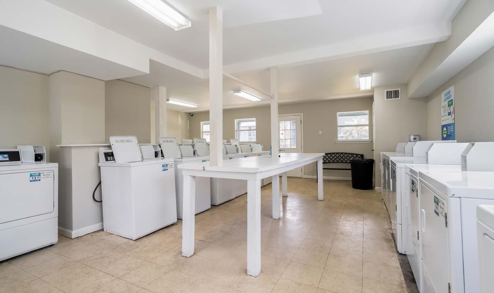Laundry room with rows of white washers and dryers around a long folding table, bright and clean.