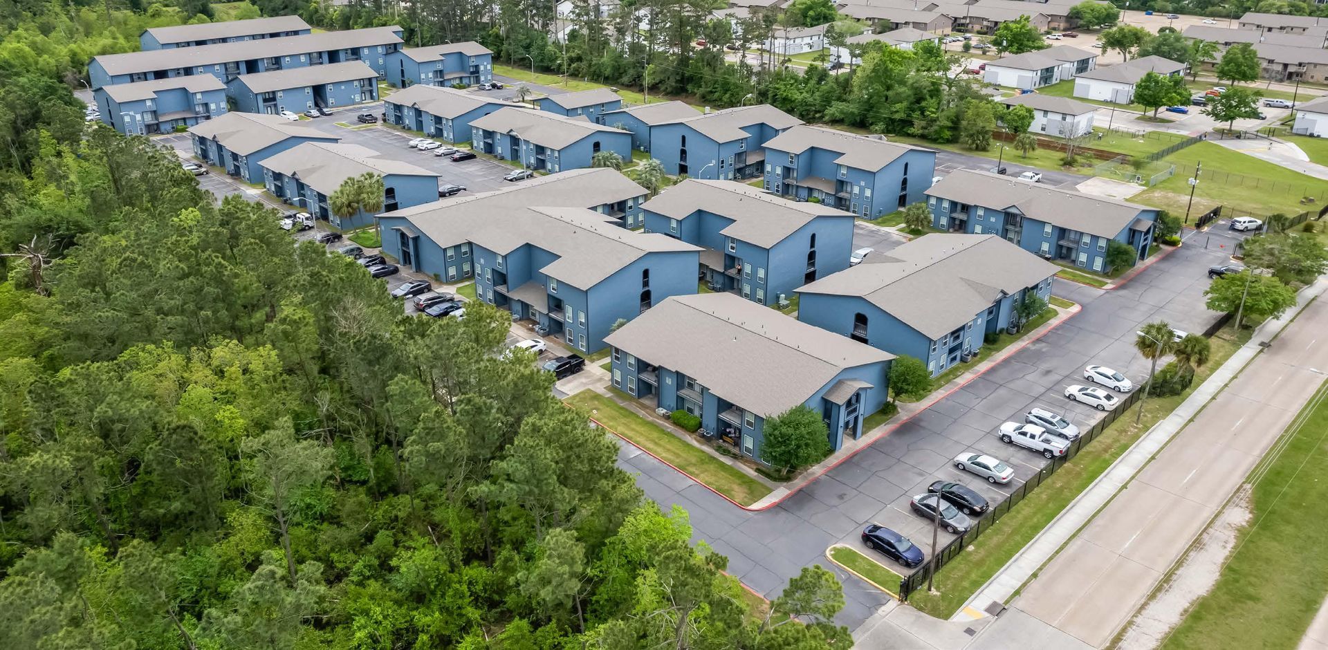 Aerial view of a blue-roofed apartment complex with parking lots and surrounding trees