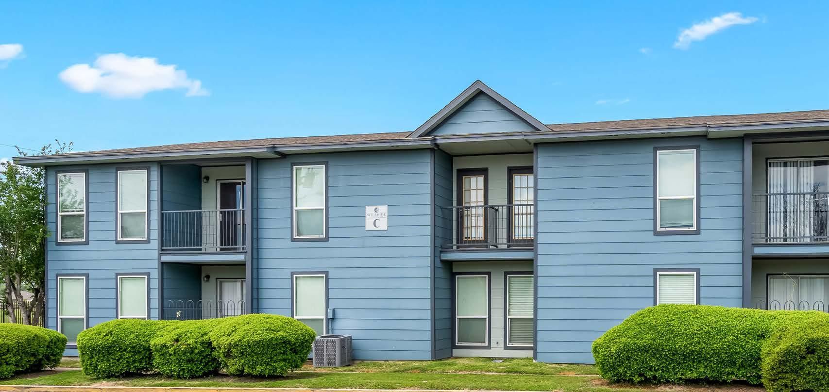Blue two-story apartment building with balconies and trimmed bushes in front