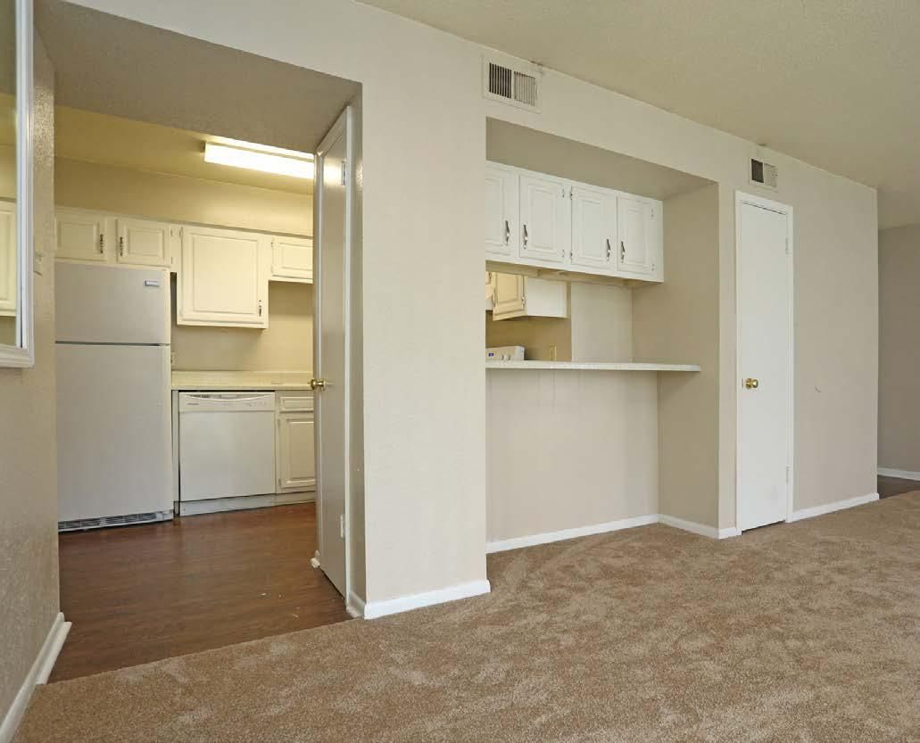 Empty apartment living area with beige carpet, open kitchen, and white walls.