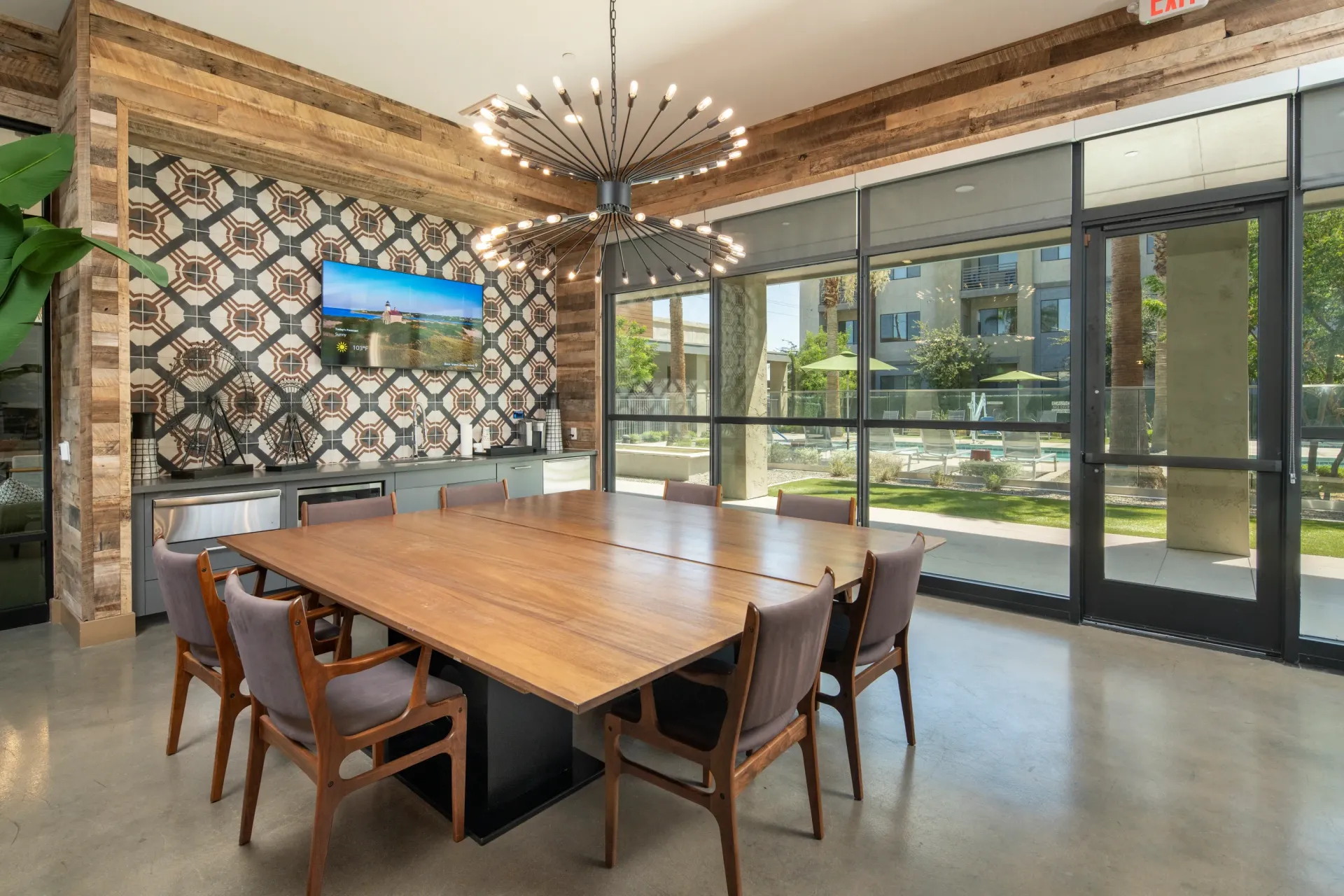 Community lounge with large wooden table, chairs, TV, and modern chandelier. Large windows overlook pool area at Parc Lofts in Tolleson, AZ.