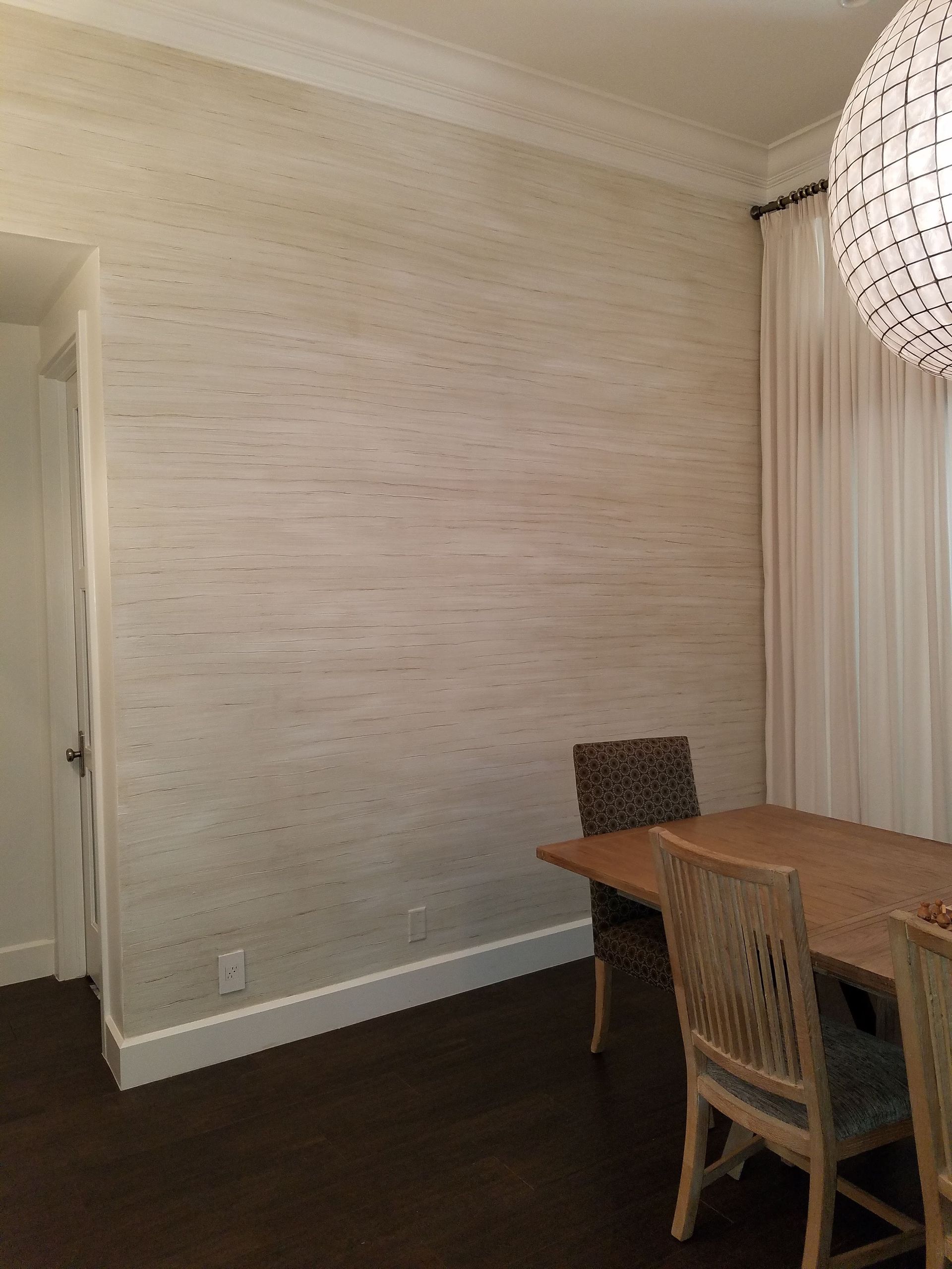 Dining room with textured beige wall, wooden table and chairs, dark floor, and white curtains.