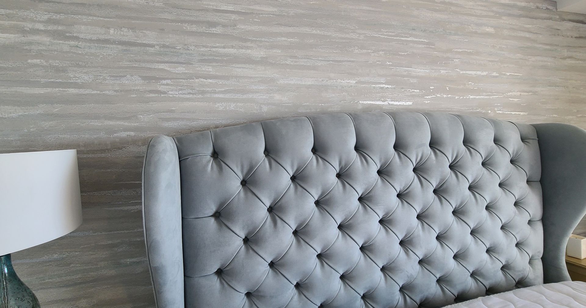 Tufted blue headboard against textured gray wall, with a white lamp base on the left.
