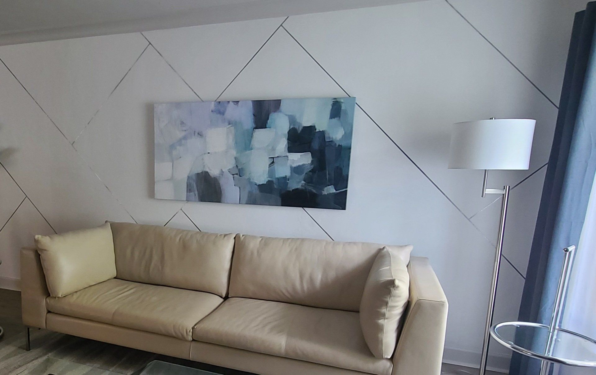 Beige sofa under an abstract painting on a geometric patterned white wall. A floor lamp stands beside a window.