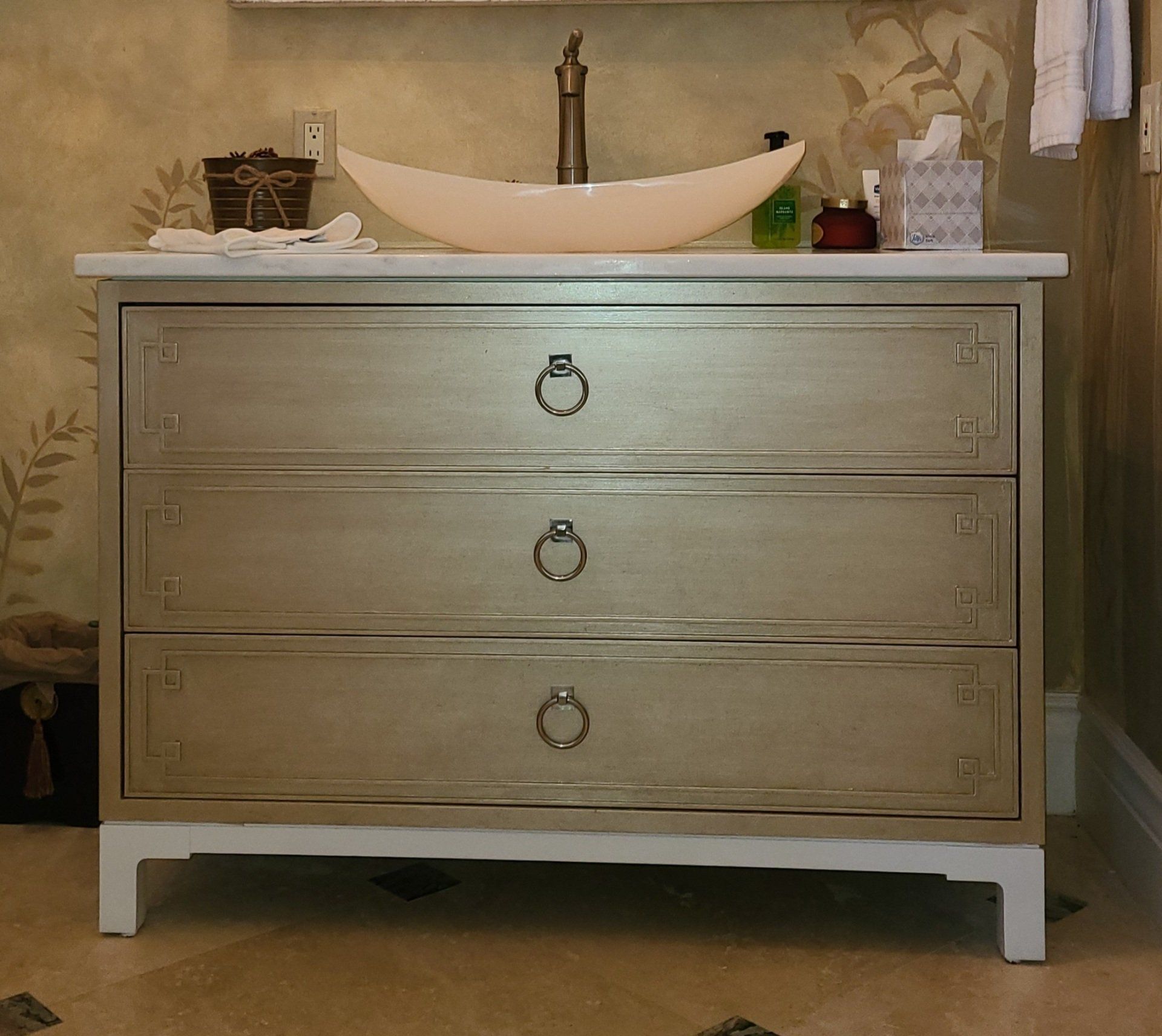 Vanity with a vessel sink. Three drawers, silver finish, ring pulls, set against a floral wall.