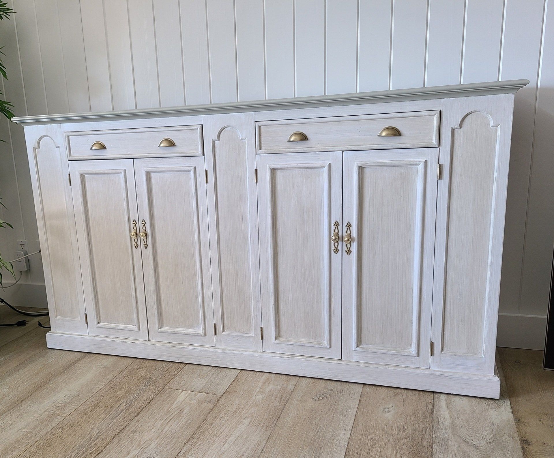 White wooden cabinet with four doors and two drawers, brass handles, against a white wall.
