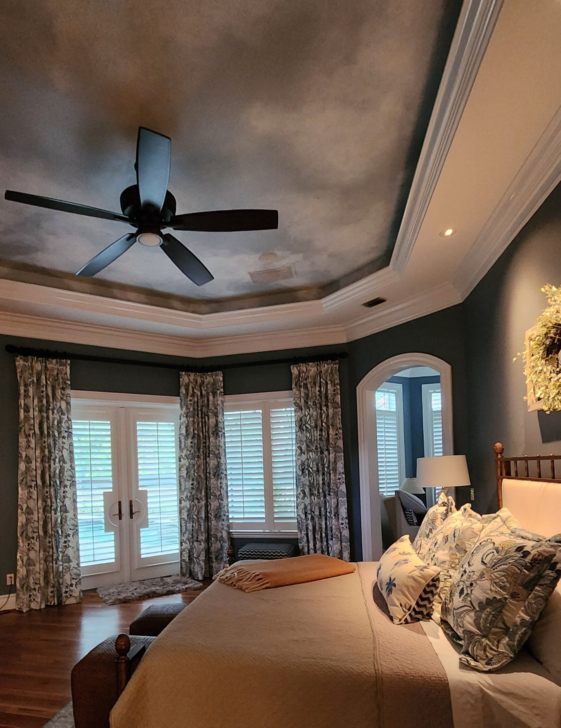 Bedroom with dark ceiling, ceiling fan, bed, blue walls, and window with patterned curtains.