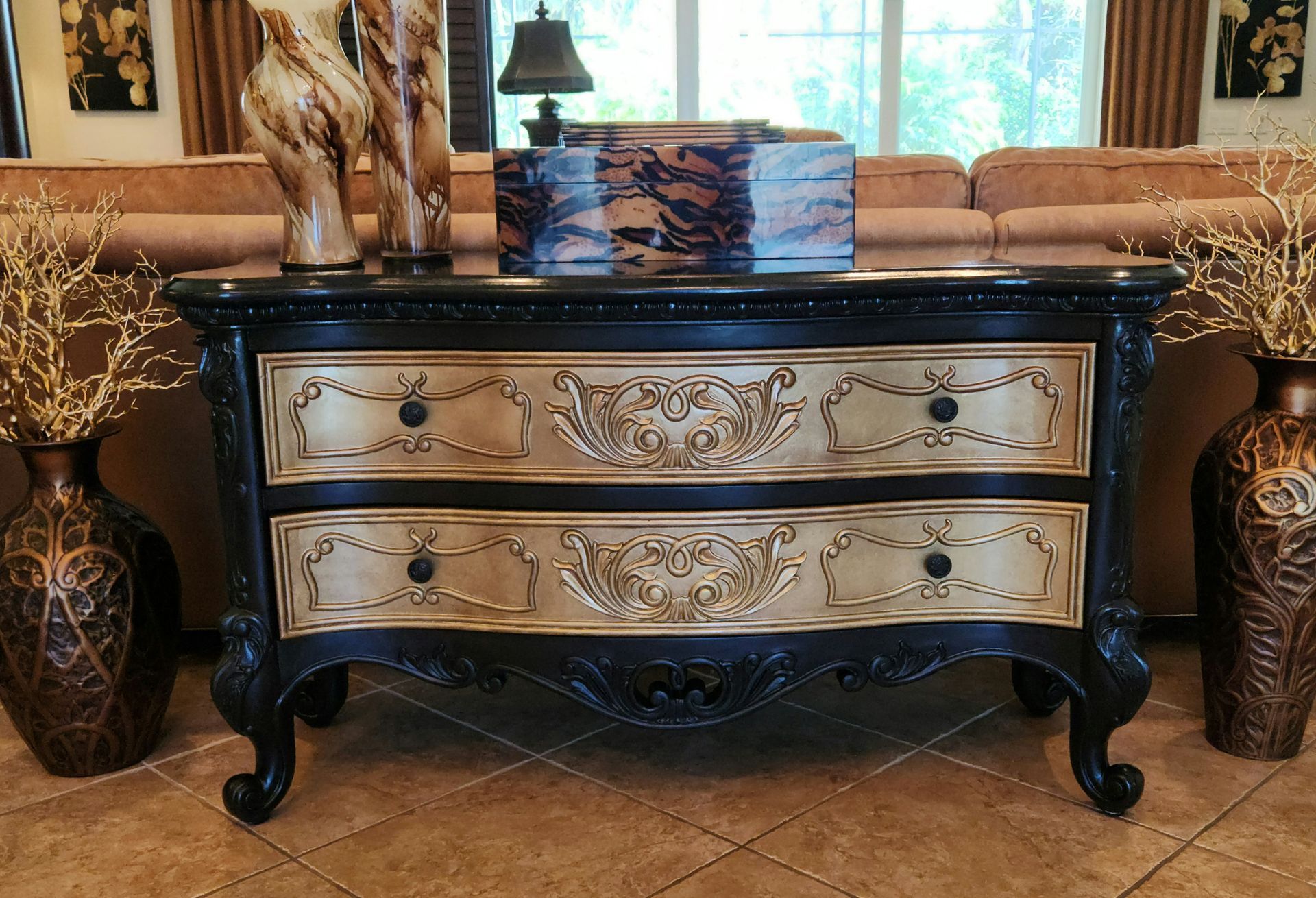 Black and gold ornate chest of drawers with two drawers, vases, and couch in the background.