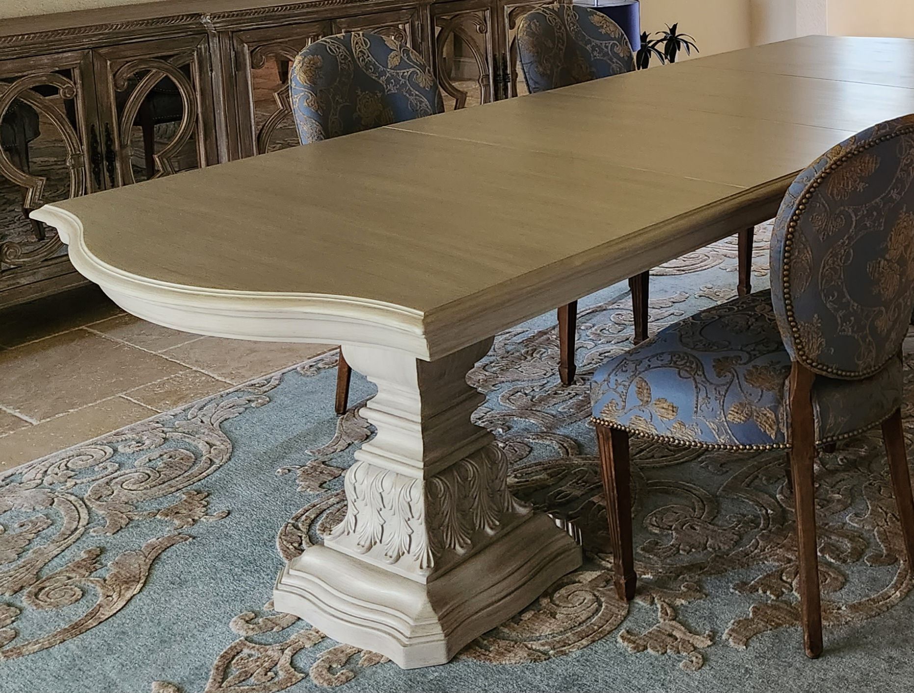 Dining table with ornate base, set with chairs on patterned rug.