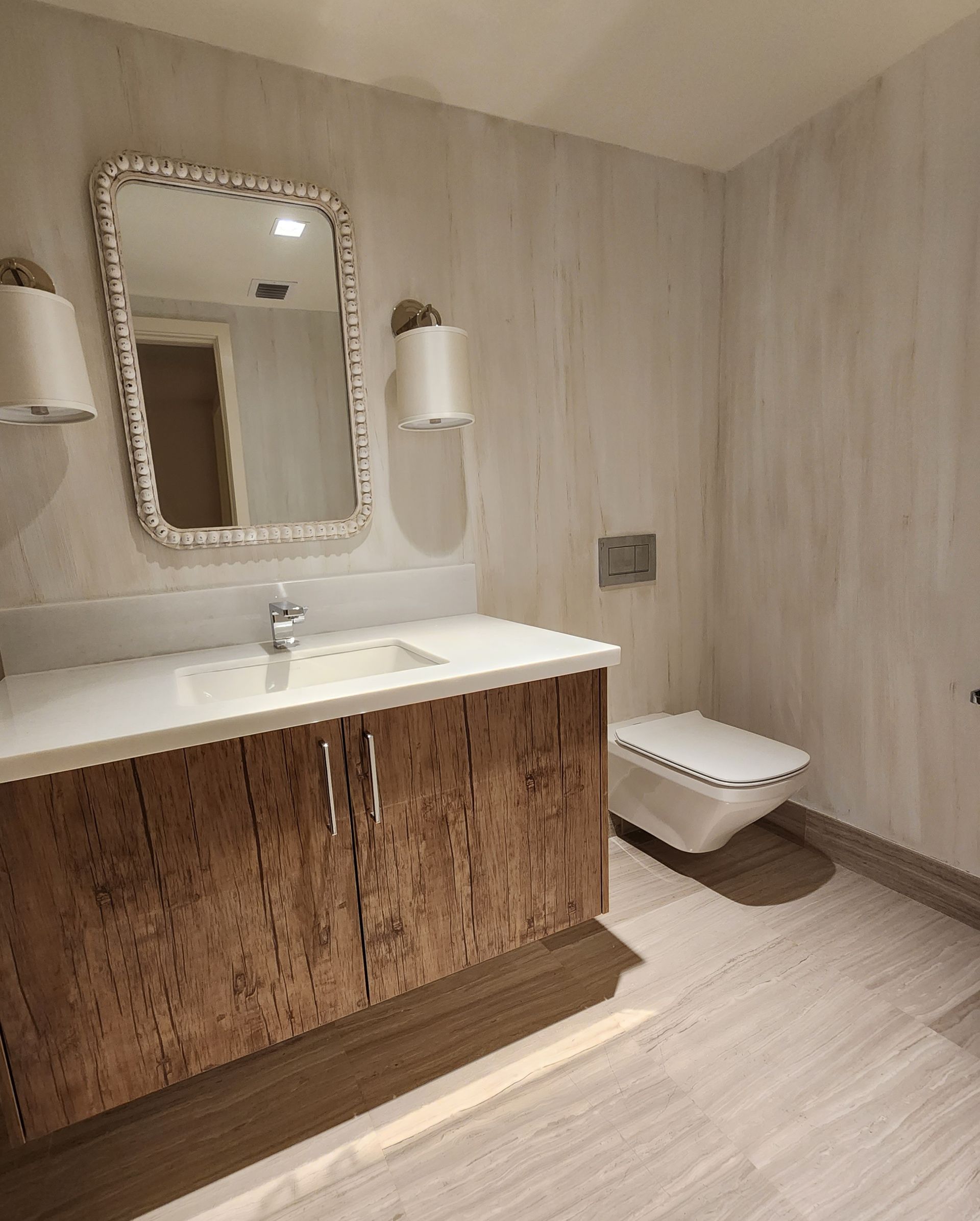 Bathroom with a wooden vanity, white countertop, toilet, and mirror with sconces on a light wood-grain wall.