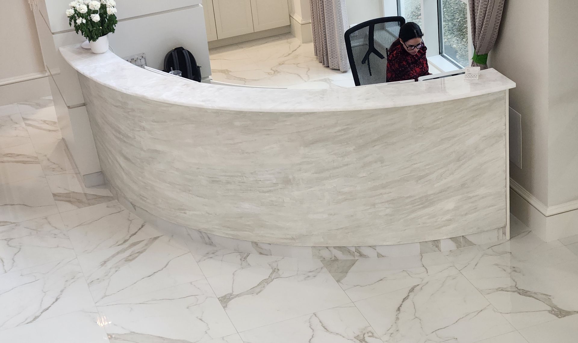 Reception desk in a light-colored office with a person sitting behind it, with marble-like flooring.