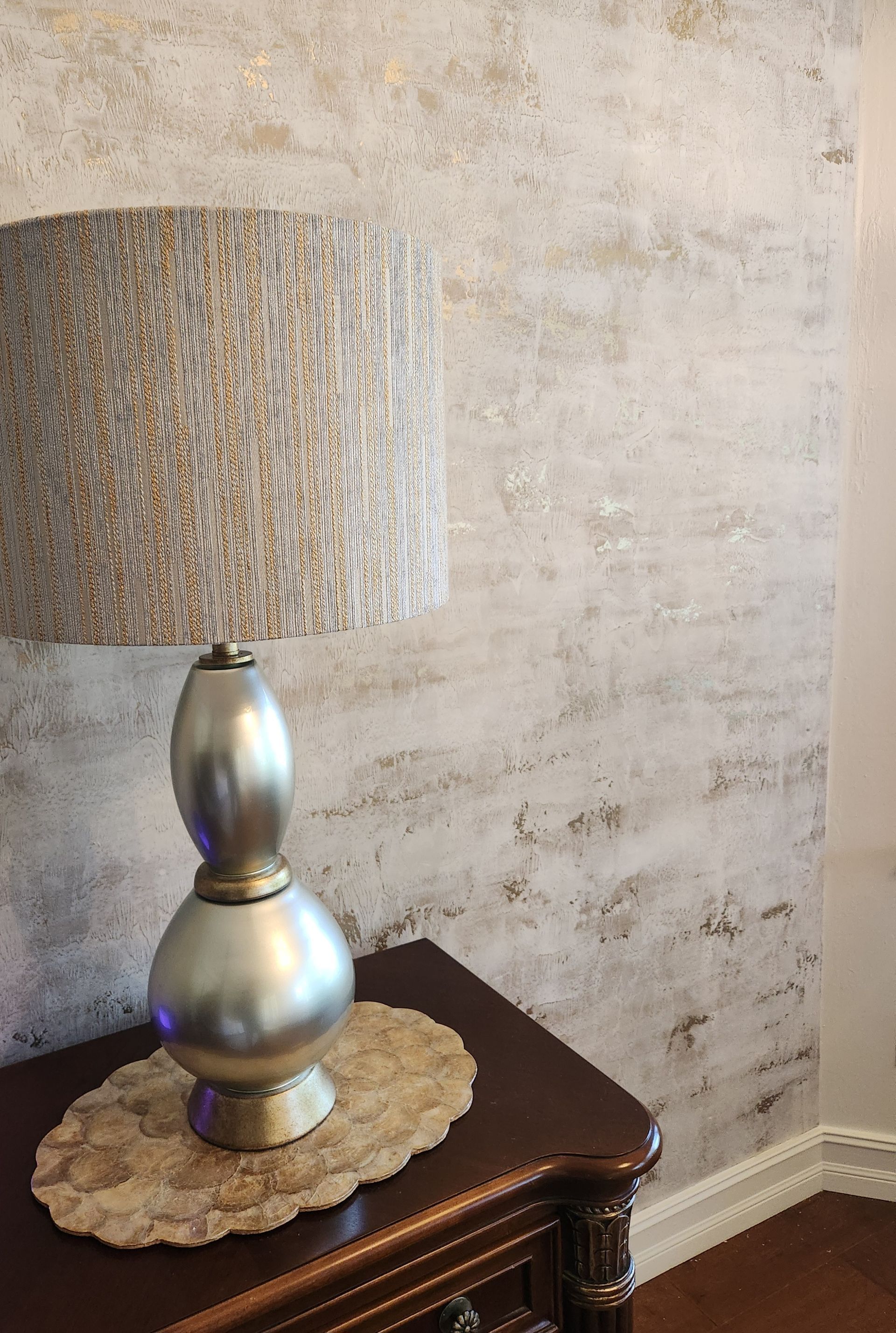 Lamp on a nightstand with textured wall. Silver lamp base, patterned lampshade, beige wall.