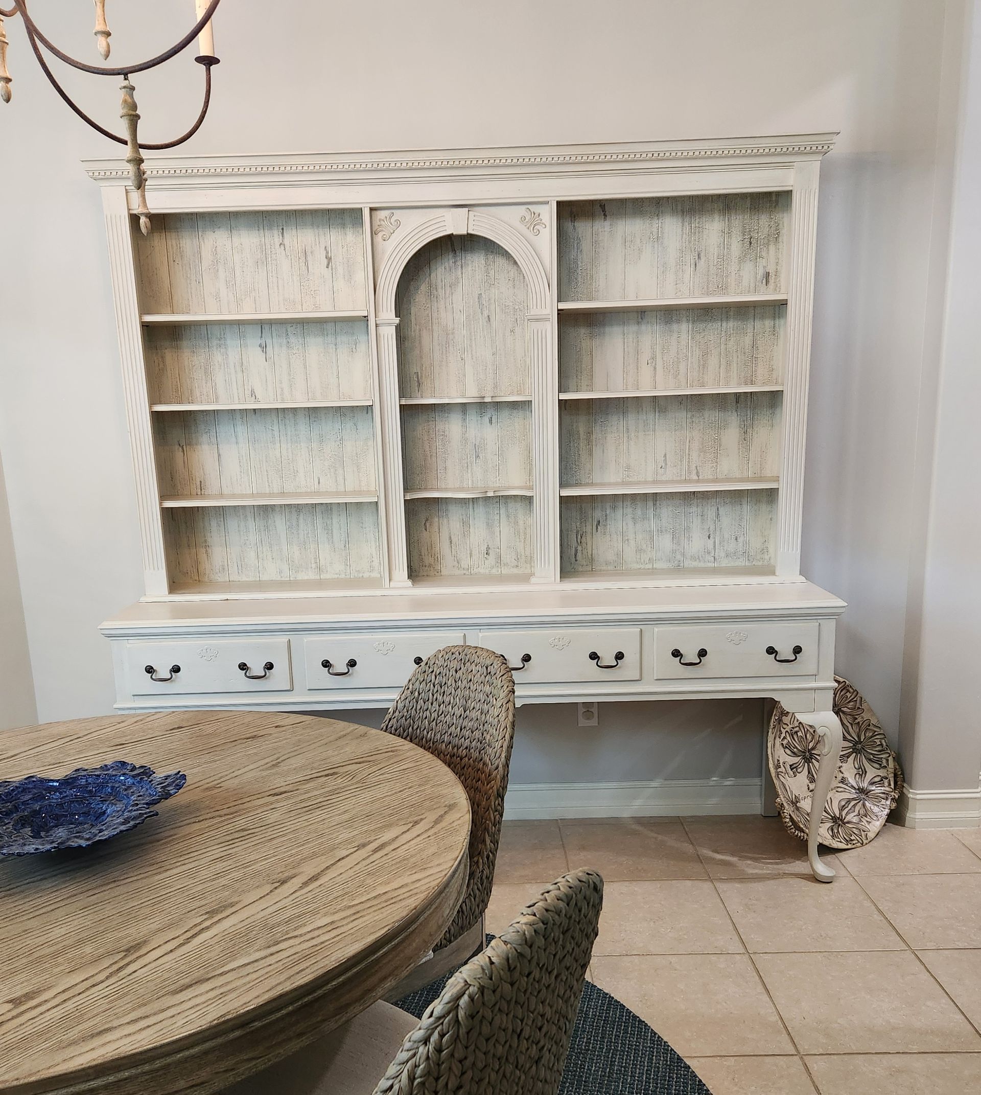 White distressed bookshelf with drawers, a round table with a blue bowl, and textured chairs.