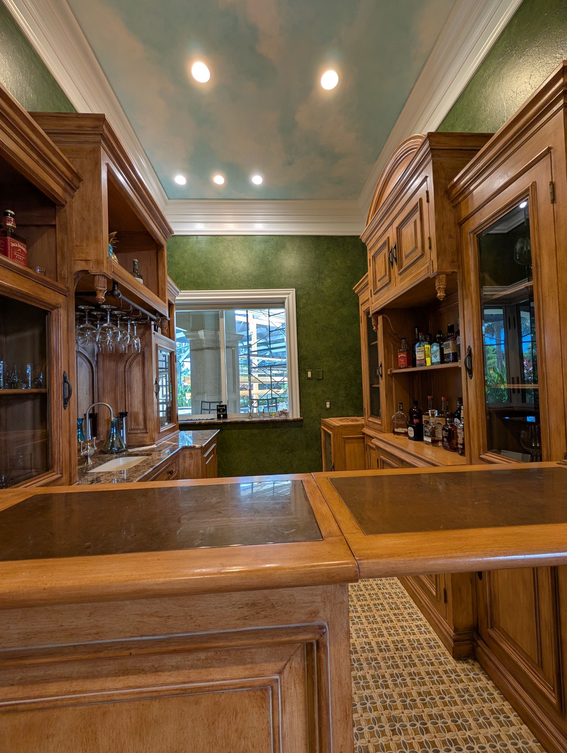 Wooden bar with liquor bottles, cabinets, and a cloudy ceiling. Window and patterned tile floor are visible.