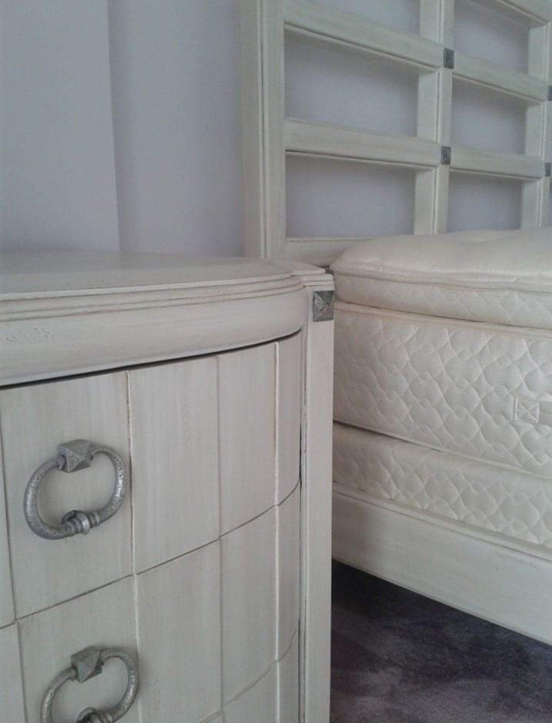 Cream-colored nightstand with silver handles next to a bed frame and mattress.