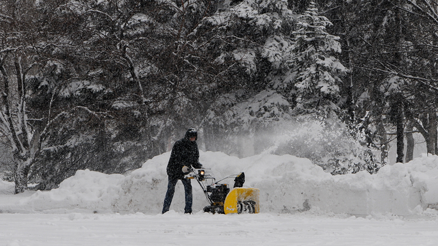 Person clears snow with a yellow snow blower in a snowy yard near trees.