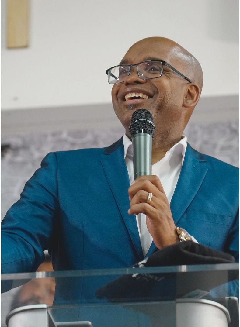 A man in a suit and glasses is sitting with his hands folded and smiling.