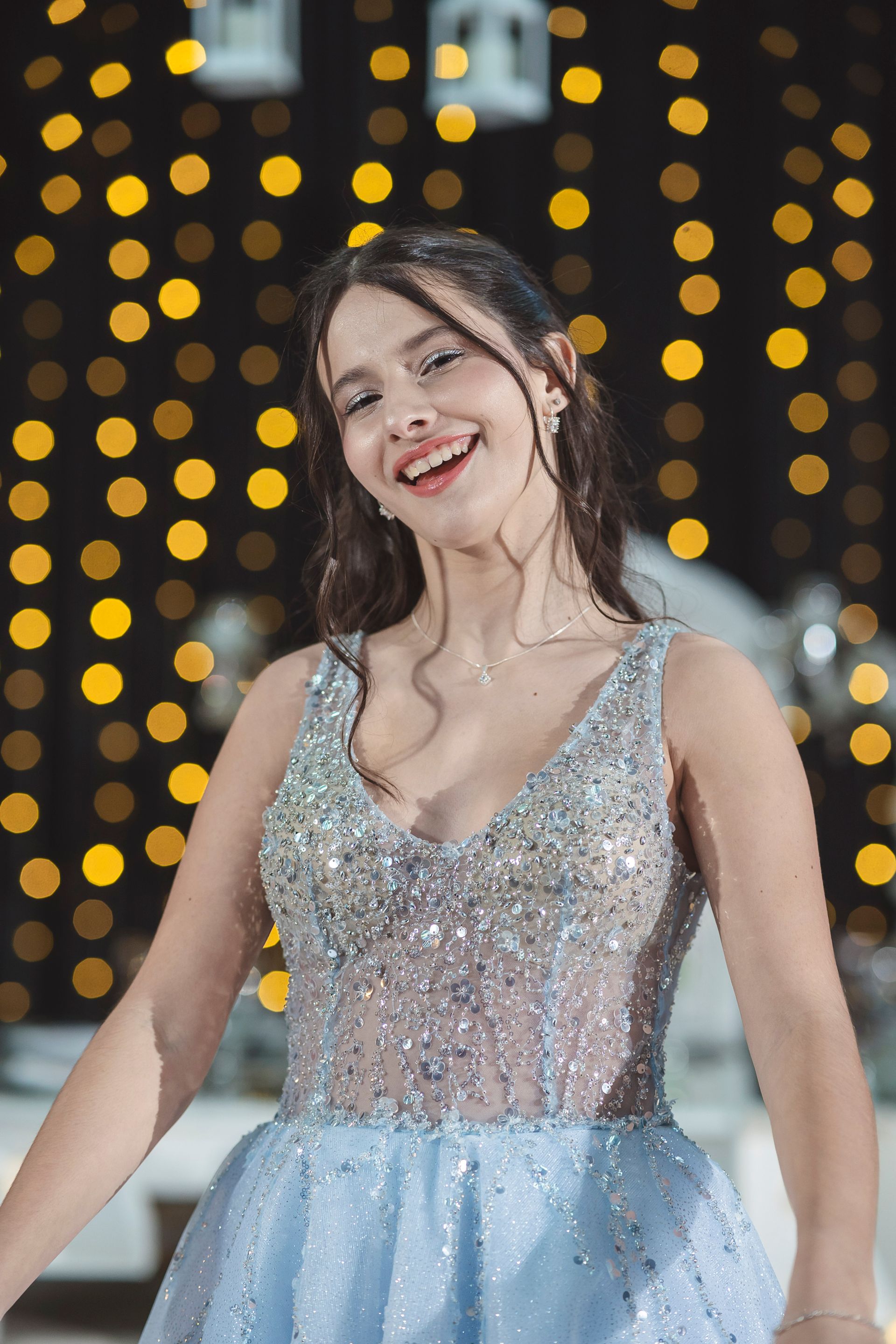 Mujer con vestido plateado y falda azul claro posando en un salón de eventos decorado con guirnaldas de luces.