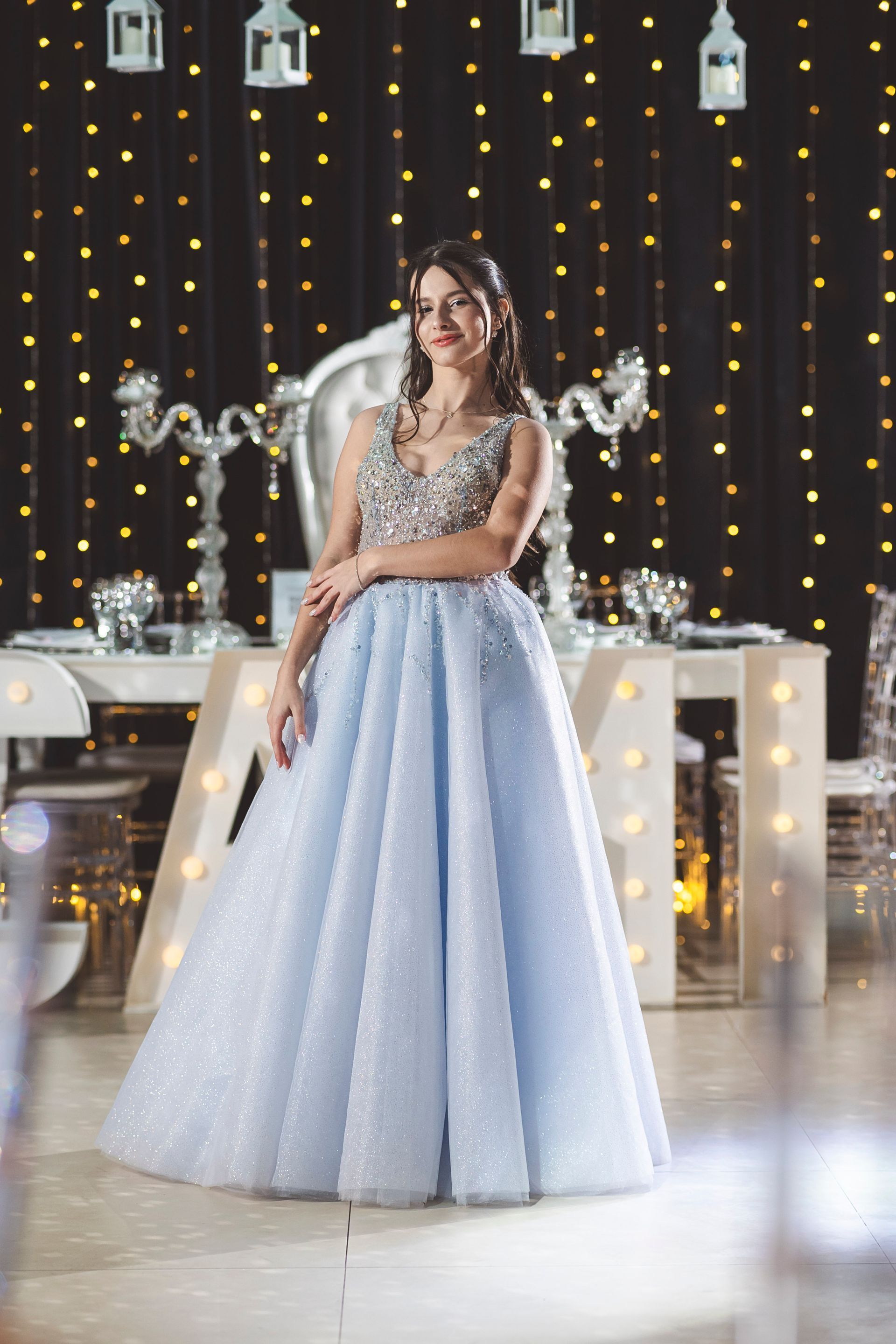 Mujer con vestido plateado y falda azul claro posando en un salón de eventos decorado con guirnaldas de luces.
