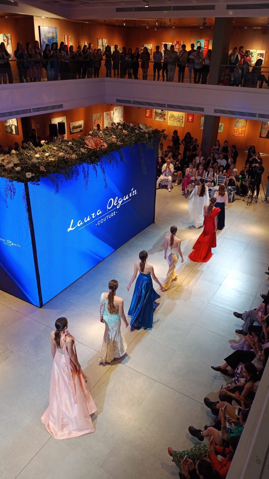 Desfile de moda en un gran salón, con modelos vestidas con vestidos en tonos pastel y rojos desfilando