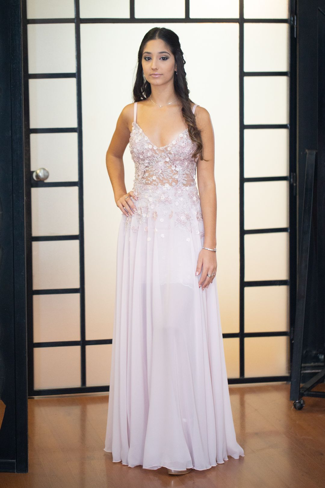 Persona con un vestido rosa pálido adornado, de pie frente a un fondo estilo shoji.