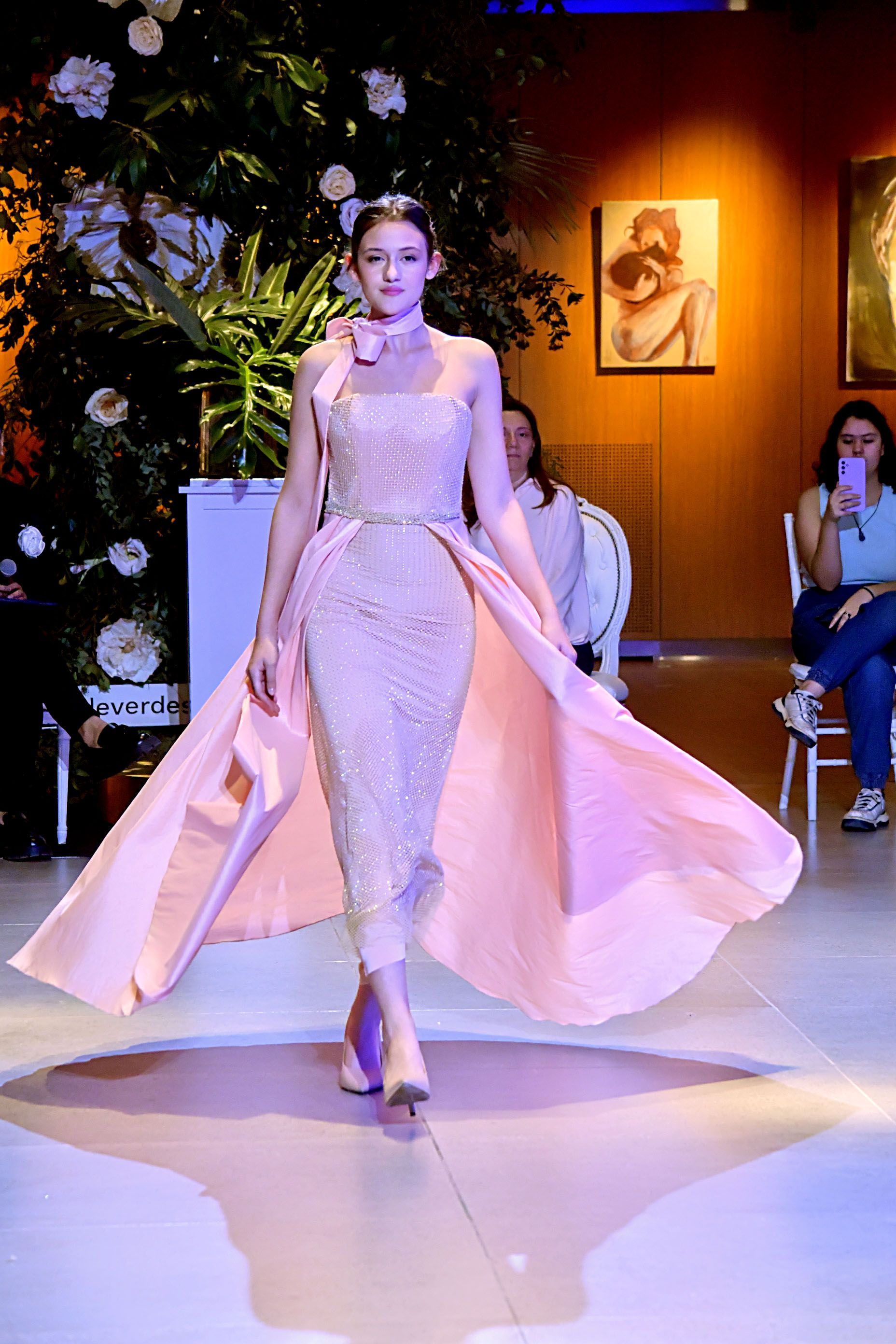 Desfile de moda en un gran salón, con modelos vestidas con vestidos en tonos pastel y rojos desfilando