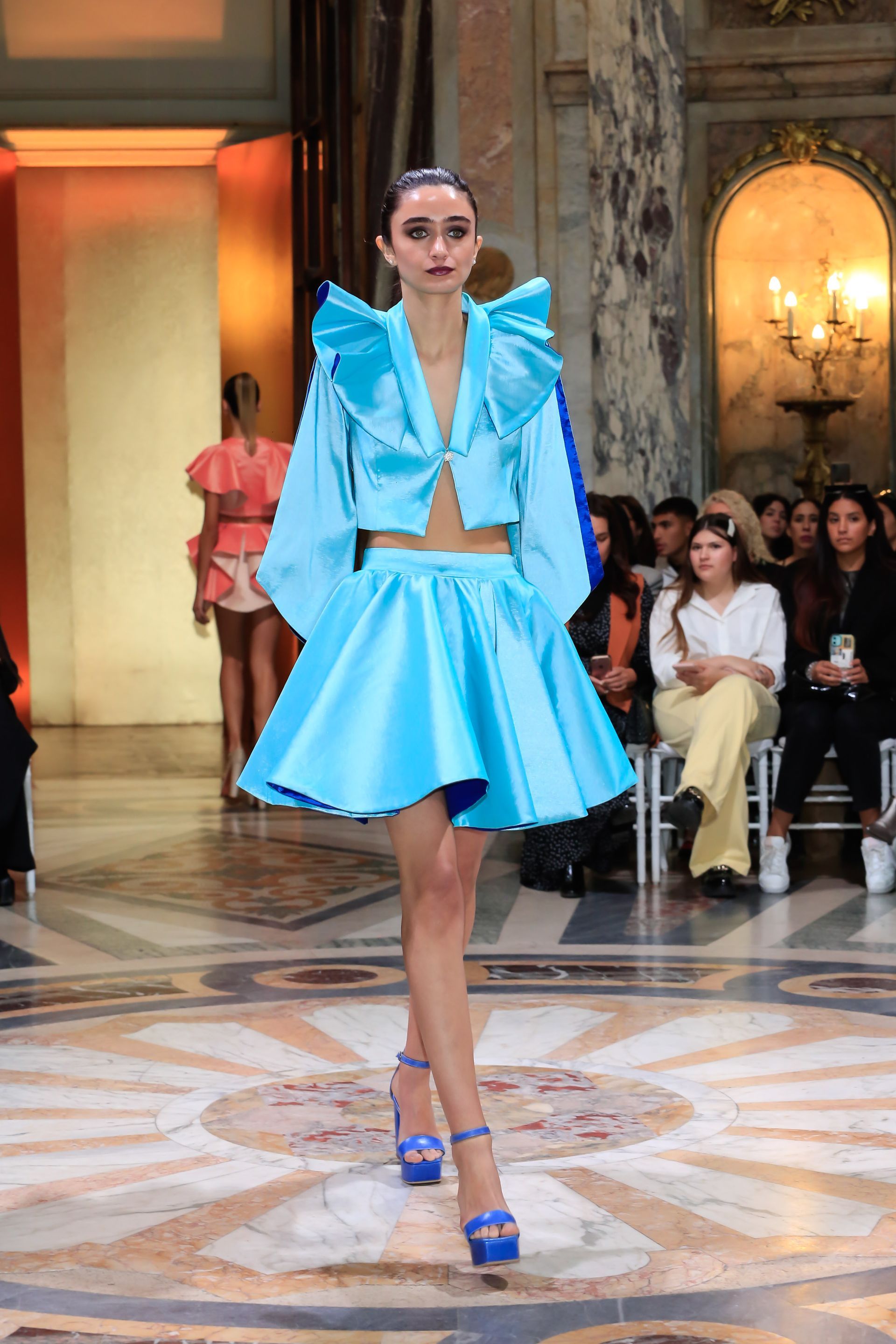 Modelos con coloridos vestidos desfilan por una pasarela en un salón de baile ornamentado, con público sentado a los lados.