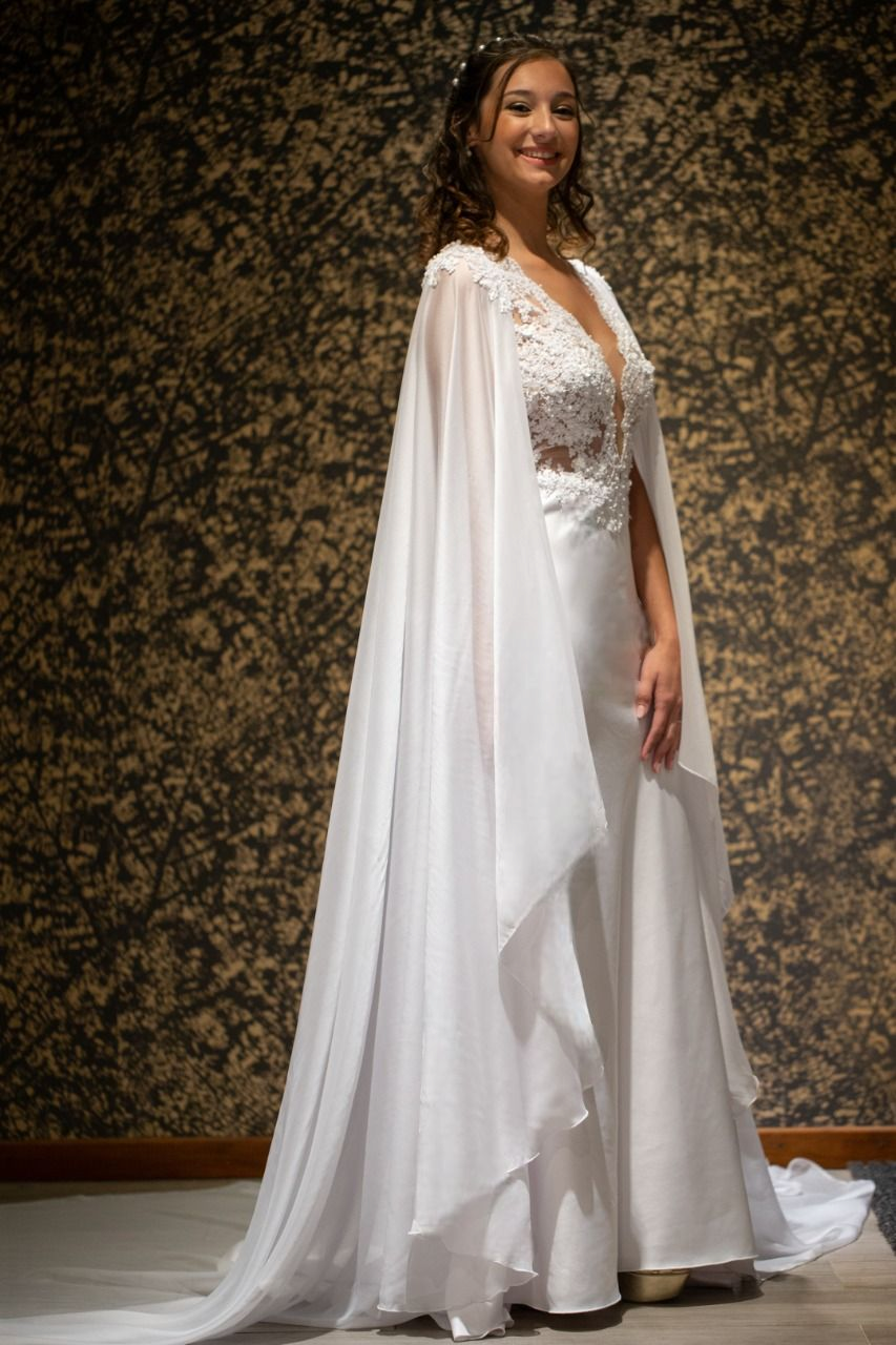 Una mujer con un vestido de novia blanco y una capa está parada frente a una pared.