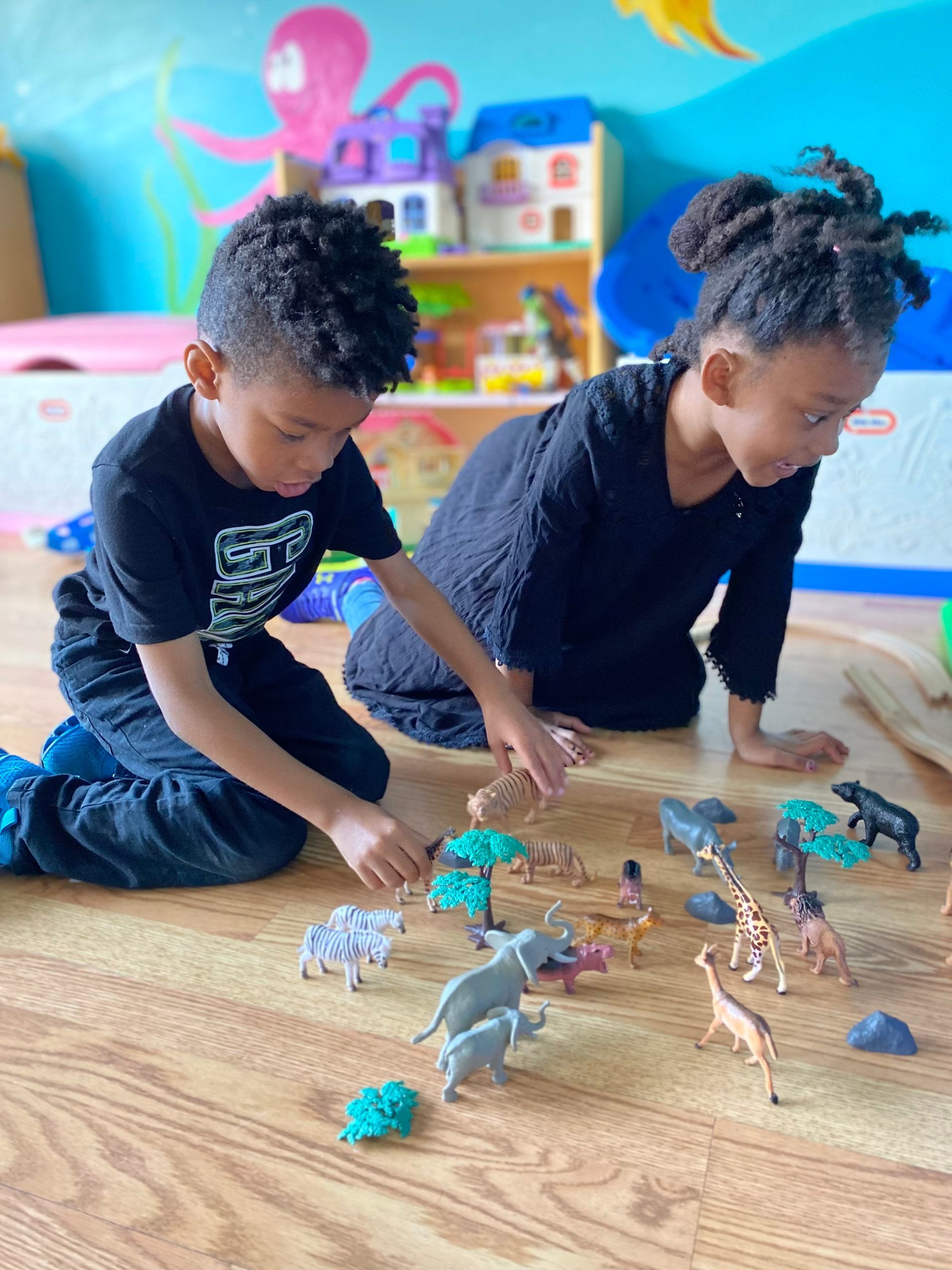 Two children, a boy and a girl, play with toy animals on a wooden floor.