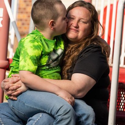 Boy kissing a woman on the cheek while sitting on her lap outside; green, blue, and black clothing.