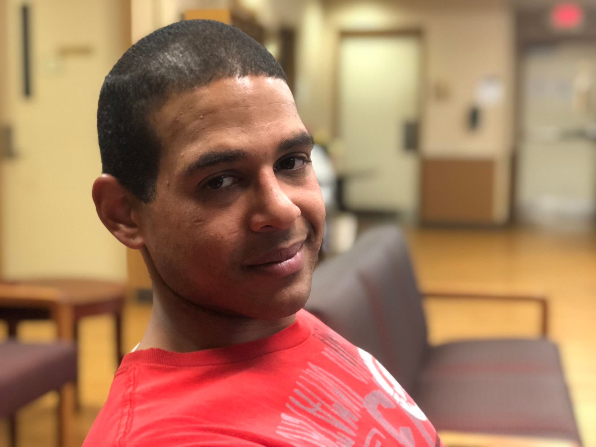 Man in red shirt smiles at camera, sitting on a sofa in a hallway.