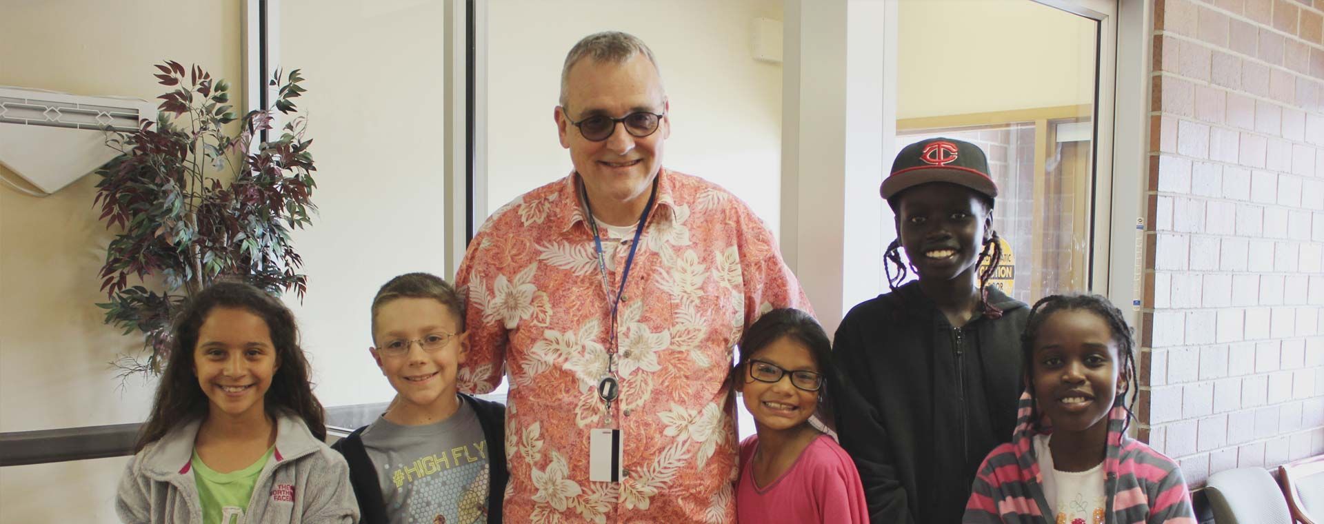 A group of children and a man smiling in a bright hallway. The man is wearing sunglasses and a floral shirt.