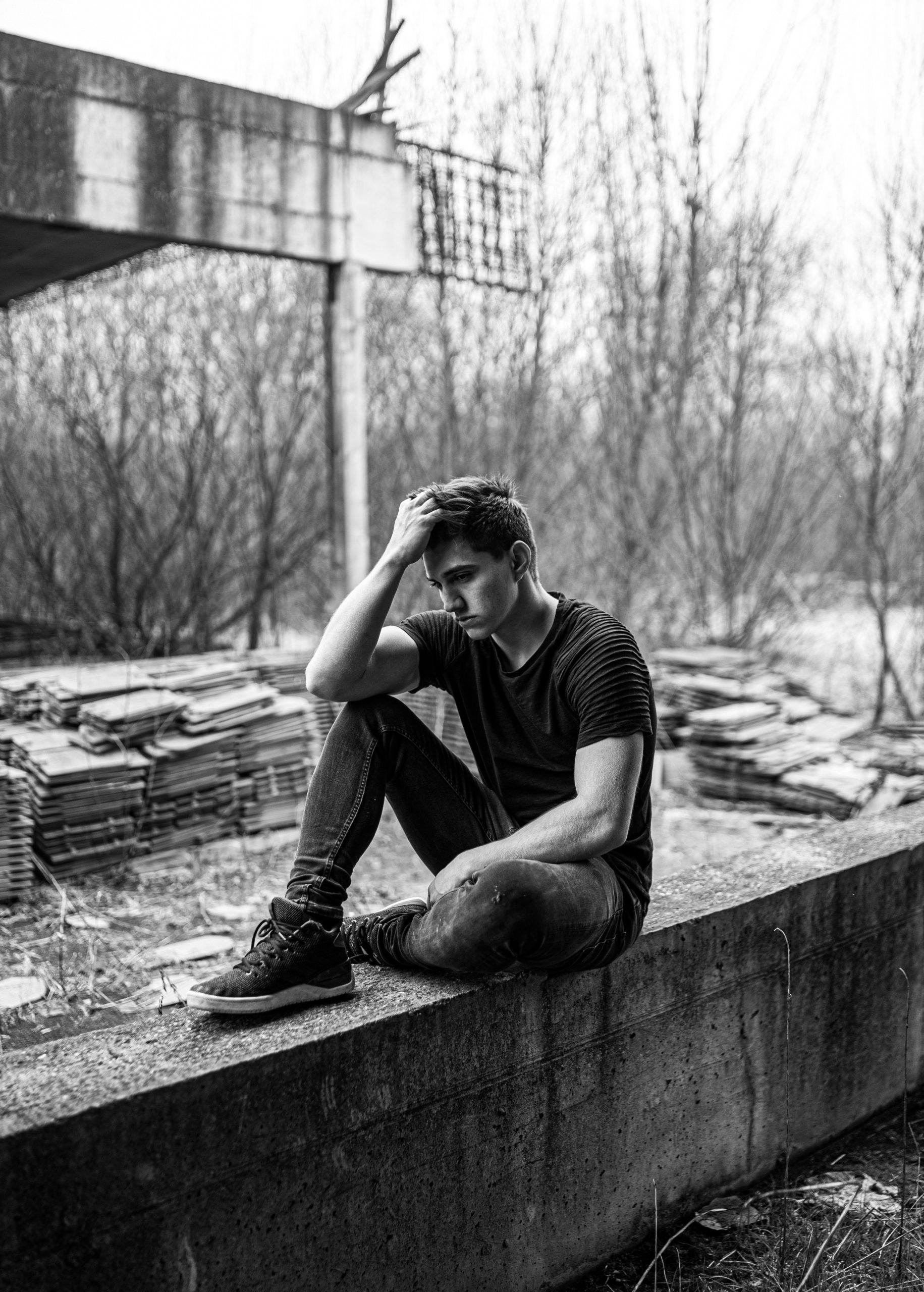 Man sits on concrete, head in hand, looking down, in desolate setting.