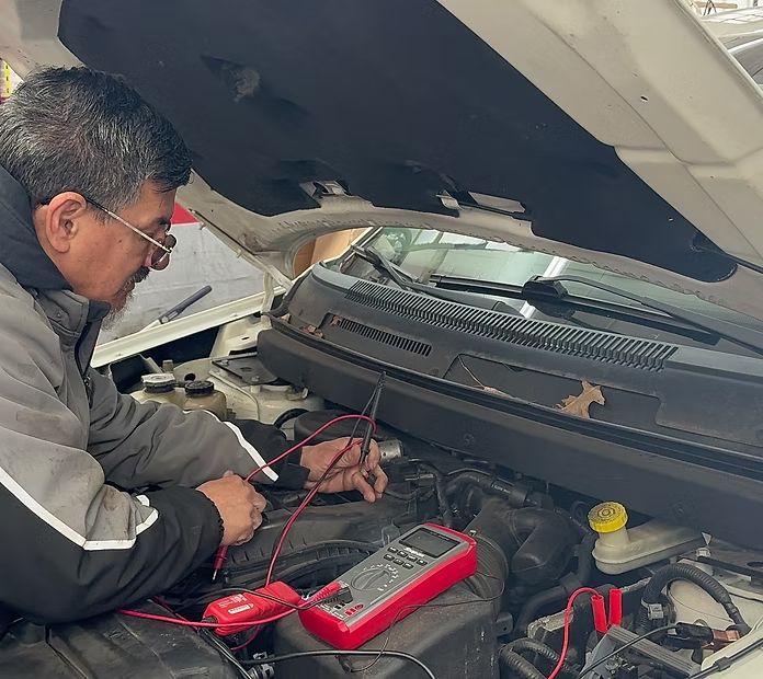 A technician in a gray jacket uses a red digital multimeter to test components under the open hood of a car. | New Pegasus Automotive