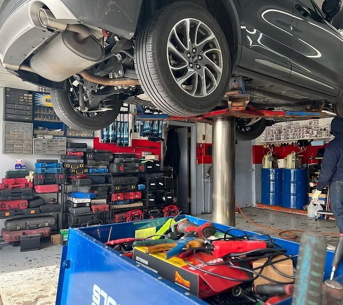 A car raised on a hydraulic lift in an auto repair shop, with a tool cart in the foreground and shelves of tools behind. | New Pegasus Automotive