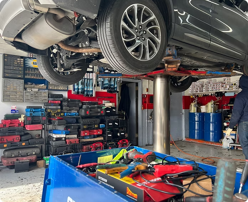 Car raised on a lift in an auto repair shop, with tools, parts, and storage shelves visible. | New Pegasus Automotive