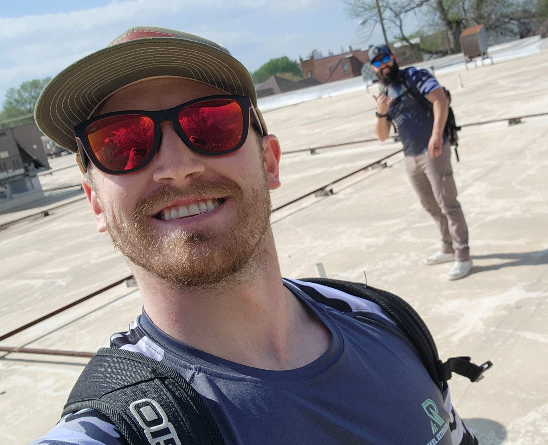 Man with red sunglasses smiles, takes selfie on rooftop. Another man stands in background.