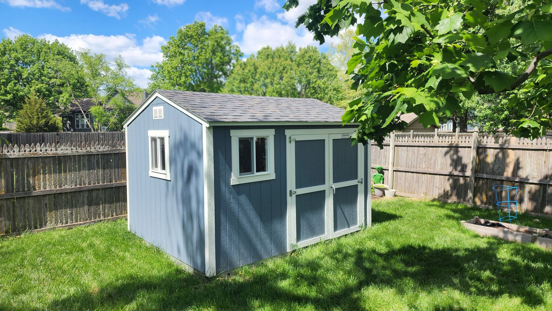 Blue shed in a grassy backyard with a wooden fence and trees on a sunny day.