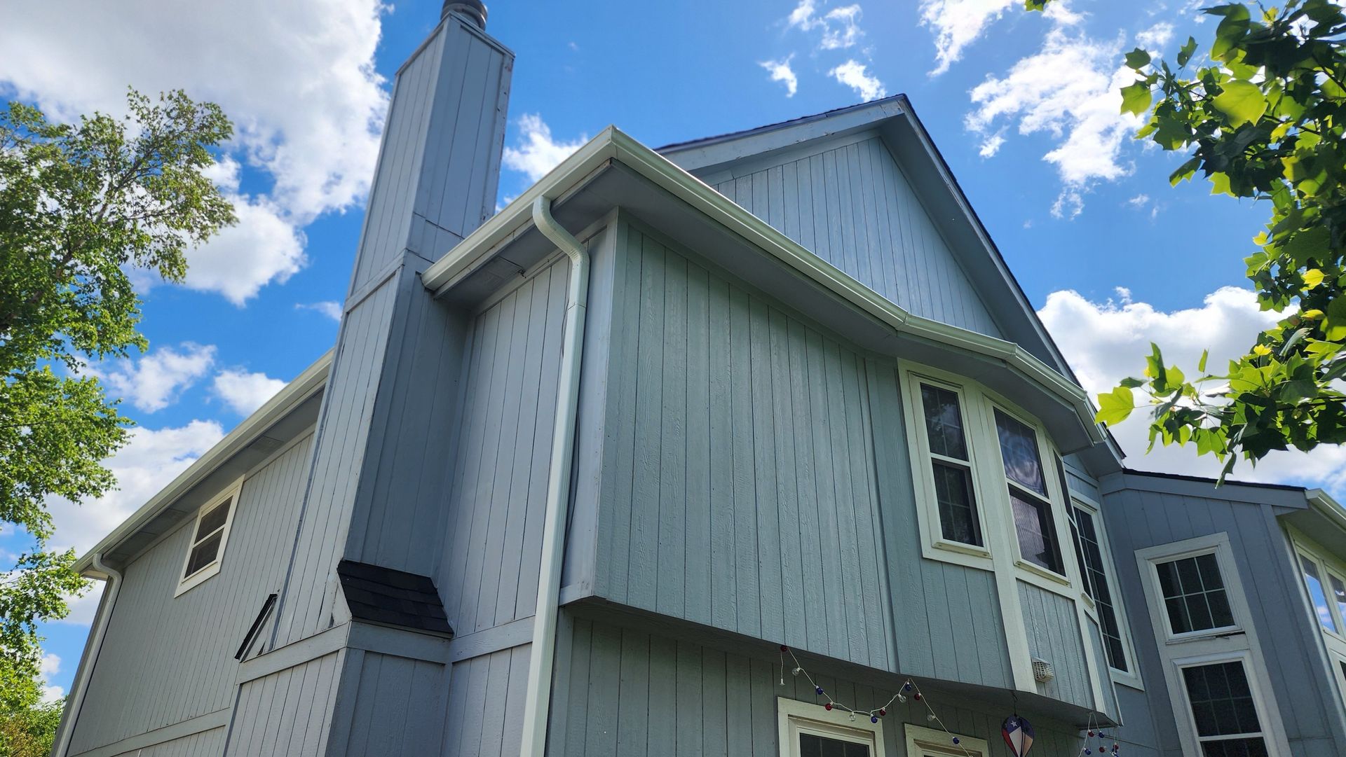 Blue-sided house with chimney against a partly cloudy sky, visible foliage on the right.
